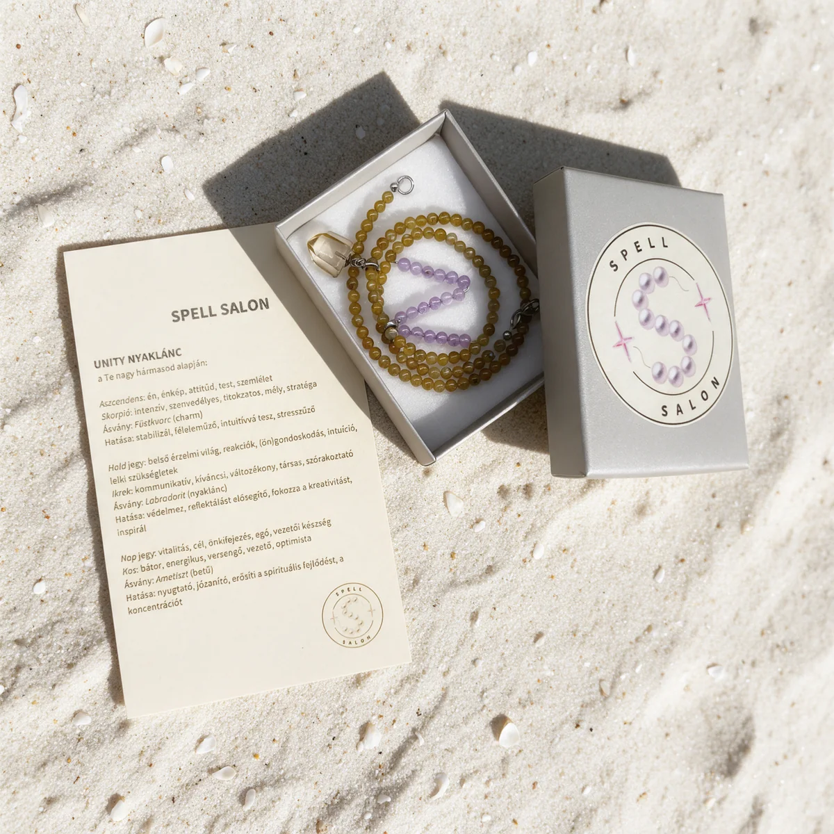 A Spell Salon necklace in its grey gift box on sand, next to a printed card explaining the three stones chosen from the birth chart